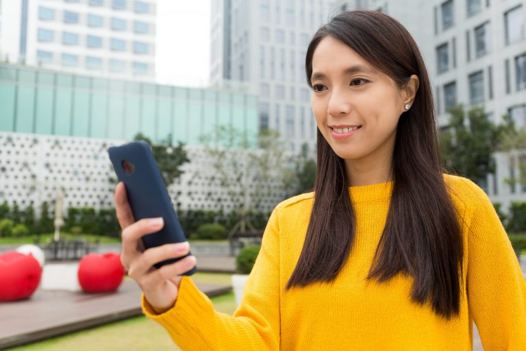 Smiling woman outdoors using her smartphone, symbolising Mocean SMS Messaging Campaign Web Portal for simple and effective customer communication in Indonesia.